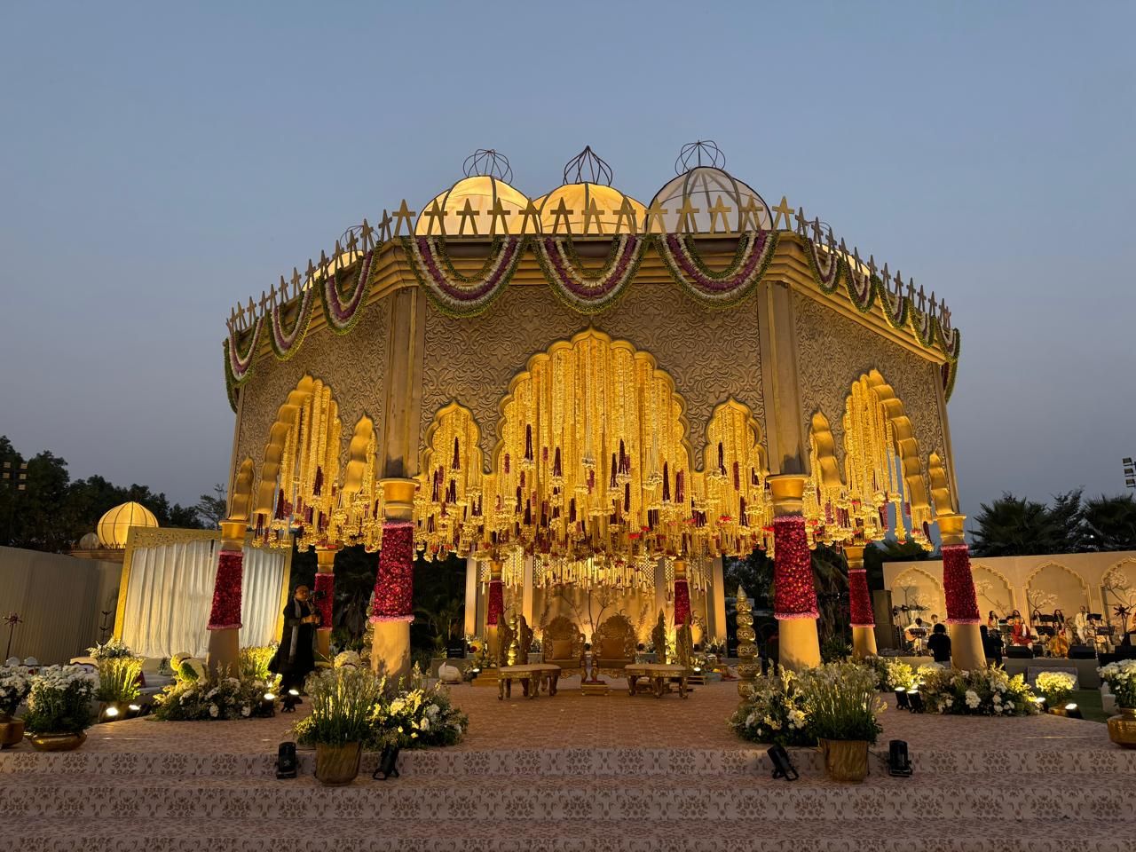 Gold mandap with hanging florals and red rose pillars at dusk